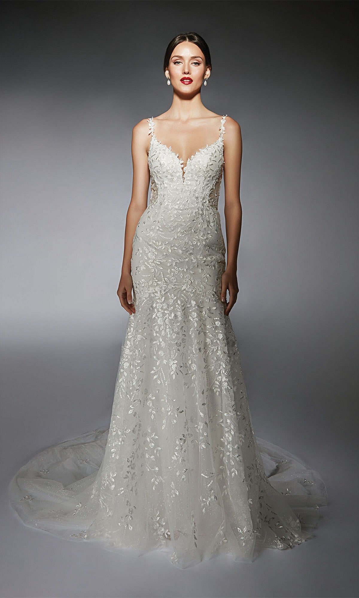 A woman wears the Alyce 7122, a long ivory dress featuring floral lace appliqués, thin straps, and a flowing train.